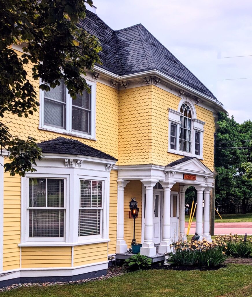 The southern facade of Le Griffon Bed & Breakfast faces Main Street. The Victorian Italianate villa is painted in Arles yellow and white. It has a mansard roof, vaulted windows, and large columns support the second floor bump-out.