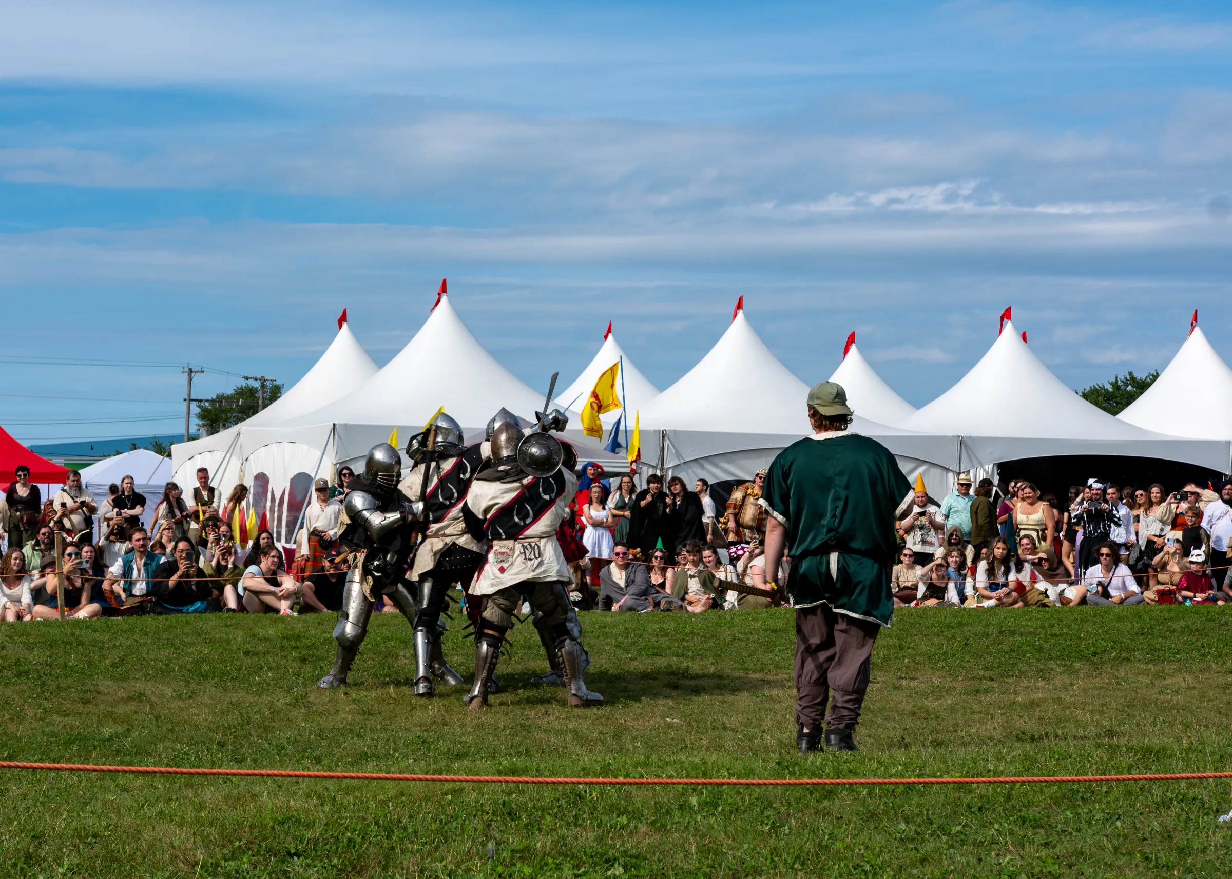 A packed crowd watches armoured fighters in a buhurt bout in front of white peaked tents at the 2026 Shediac Renaissance Faire