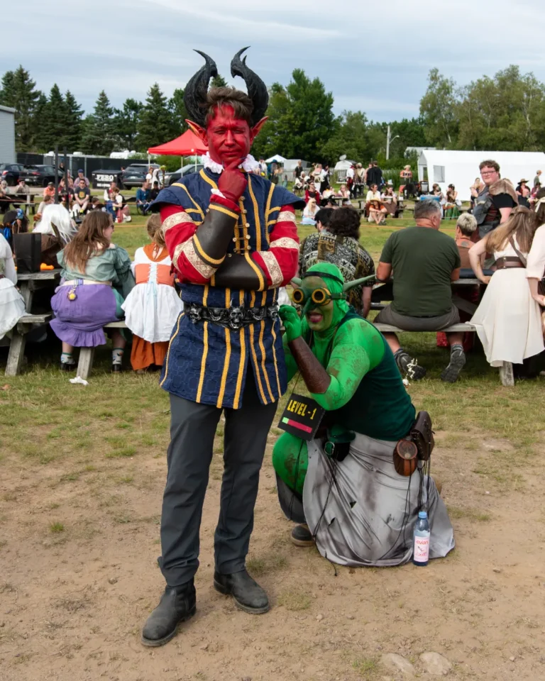 A red-faced horned demon and a green goblin pose among picnic tables at the Faire